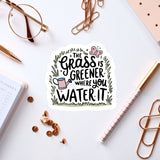a sticker with a hand-drawn illustration of a watering can surrounded by grass, along with the text "The grass is greener where you water it".