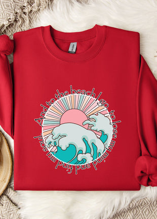 A red sweater with a graphic design of a beach scene and the text "To the beach, I love you" is displayed on a bed with a white blanket and a white mug with a snowflake design.