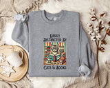A gray sweatshirt with a graphic design of a cat surrounded by books and a cup of tea, along with the text "Easily Distracted By Cats & Books".