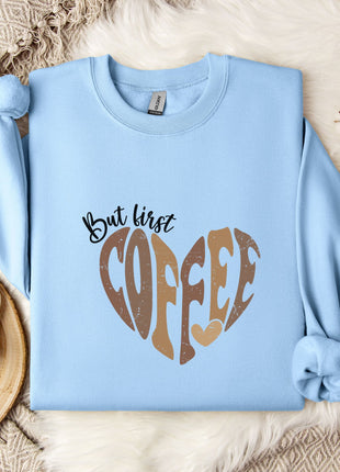 A light blue sweatshirt with a heart-shaped design and the text "But first coffee" printed on it, along with a mug and some flowers on a bed.