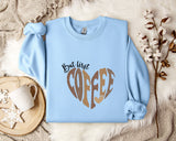 A light blue sweatshirt with a heart-shaped design and the text "But first coffee" printed on it, along with a mug and some flowers on a bed.