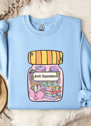 A light blue sweatshirt with a colorful graphic of a jar filled with various objects, including books and flowers, is displayed on a bed with a white blanket and a mug.