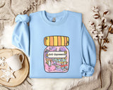 A light blue sweatshirt with a colorful graphic of a jar filled with various objects, including books and flowers, is displayed on a bed with a white blanket and a mug.