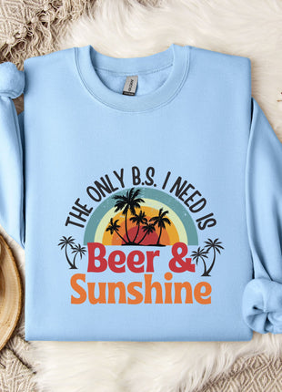 A light blue sweatshirt with a graphic design that says "The only B.S. I need is Beer & Sunshine" printed on it, along with a palm tree and sunset. The sweatshirt is laid out on a bed with a white blanket and a mug.