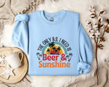 A light blue sweatshirt with a graphic design that says "The only B.S. I need is Beer & Sunshine" printed on it, along with a palm tree and sunset. The sweatshirt is laid out on a bed with a white blanket and a mug.