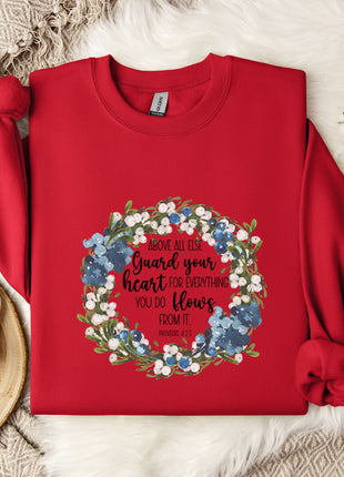 A red sweater with a floral wreath design and a quote printed on it, along with a mug and some white flowers, all placed on a white fur rug.