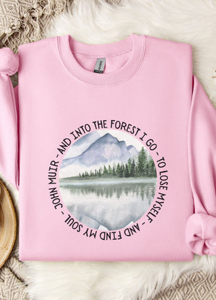 A pink sweatshirt with a mountain landscape design and a quote around the edge, placed on a white fur rug with a mug and a branch of cotton bolls nearby.