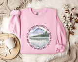 A pink sweatshirt with a mountain landscape design and a quote around the edge, placed on a white fur rug with a mug and a branch of cotton bolls nearby.