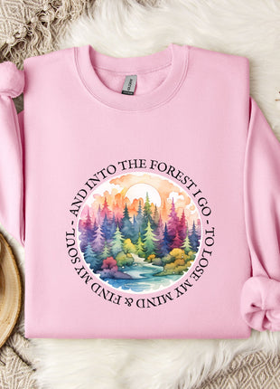 A pink sweatshirt with a colorful forest scene and the text "AND INTO THE FOREST I GO, SOIL MY SOUL AND I LOVE MINIMUM MINDS" printed on it, along with a mug and some white flowers on a bed.