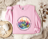 A pink sweatshirt with a colorful forest scene and the text "AND INTO THE FOREST I GO, SOIL MY SOUL AND I LOVE MINIMUM MINDS" printed on it, along with a mug and some white flowers on a bed.