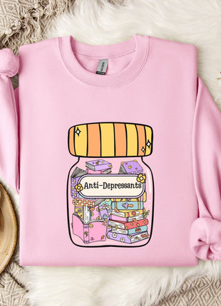 A pink sweatshirt with a graphic design of a jar containing various objects, including books and a teapot, along with the text "Anti-Depressants". The sweatshirt is laid out on a white fur rug, with a mug and a branch of cotton bolls nearby.