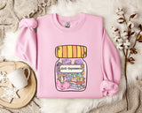 A pink sweatshirt with a graphic design of a jar containing various objects, including books and a teapot, along with the text "Anti-Depressants". The sweatshirt is laid out on a white fur rug, with a mug and a branch of cotton bolls nearby.