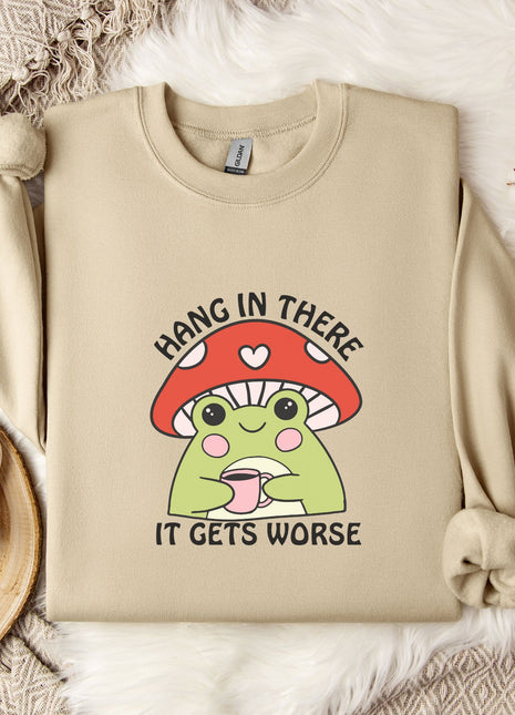 a beige sweatshirt with a mushroom graphic and the text "HANG IN THERE IT GETS WORSE" printed on it. The sweatshirt is placed on a bed with a white blanket and a mug. There are also some flowers and a snowflake in the background.