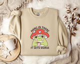 a beige sweatshirt with a mushroom graphic and the text "HANG IN THERE IT GETS WORSE" printed on it. The sweatshirt is placed on a bed with a white blanket and a mug. There are also some flowers and a snowflake in the background.