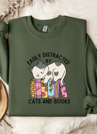 a green sweatshirt with a graphic design of a cat sitting on a stack of books, accompanied by the text "EASILY DISTracted BY CATS AND BOOKS". The sweatshirt is laid out on a bed with a white blanket, and there are some books and a mug nearby.