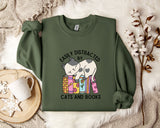 a green sweatshirt with a graphic design of a cat sitting on a stack of books, accompanied by the text "EASILY DISTracted BY CATS AND BOOKS". The sweatshirt is laid out on a bed with a white blanket, and there are some books and a mug nearby.
