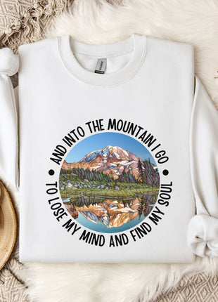 A white sweatshirt with a mountain landscape design and the text "And into the mountain I go to lose my mind and find my soul" printed on it, along with a mug and some flowers on a bed.