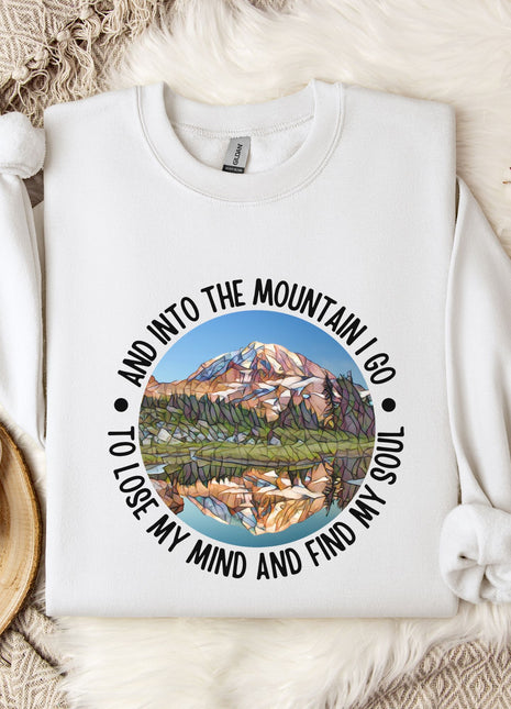 A white sweatshirt with a mountain landscape design and the text "And into the mountain I go to lose my mind and find my soul" printed on it, along with a mug and some flowers on a bed.