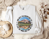 A white sweatshirt with a mountain landscape design and the text "And into the mountain I go to lose my mind and find my soul" printed on it, along with a mug and some flowers on a bed.