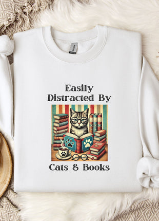 A white t-shirt with a graphic design of a cat surrounded by books, along with the text "Easily Distracted By Cats & Books". The t-shirt is placed on a bed with a white blanket and a mug.