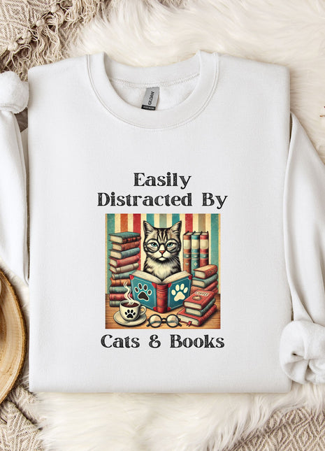 A white t-shirt with a graphic design of a cat surrounded by books, along with the text "Easily Distracted By Cats & Books". The t-shirt is placed on a bed with a white blanket and a mug.