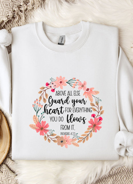 A white sweatshirt with a floral design and text printed on it, placed on a bed with a white blanket and a white mug.