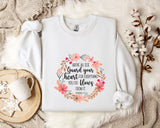 A white sweatshirt with a floral design and text printed on it, placed on a bed with a white blanket and a white mug.