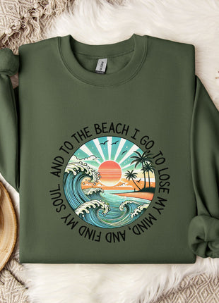 A green sweatshirt with a graphic design depicting a beach scene, along with the text "And to the beach I go, I lose my mind" printed on it. The sweatshirt is laid out on a bed with a white blanket and a white mug, and there are some white flowers nearby.