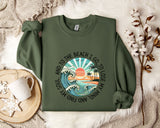 A green sweatshirt with a graphic design depicting a beach scene, along with the text "And to the beach I go, I lose my mind" printed on it. The sweatshirt is laid out on a bed with a white blanket and a white mug, and there are some white flowers nearby.