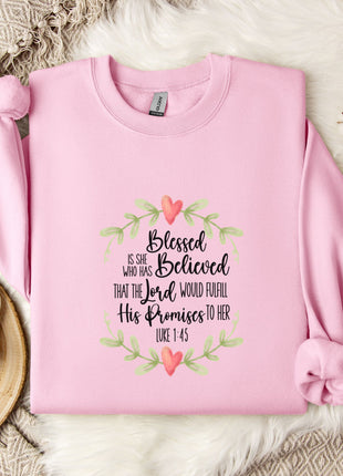 A pink sweatshirt with a printed Bible verse and a floral design, along with a white mug and a branch of cotton flowers, all placed on a white fur rug.