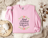 A pink sweatshirt with a printed Bible verse and a floral design, along with a white mug and a branch of cotton flowers, all placed on a white fur rug.