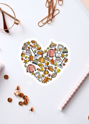 a heart-shaped sticker with various fall-themed items such as pumpkins, leaves, and a cup, surrounded by office supplies like pens, paper clips, and a notebook.