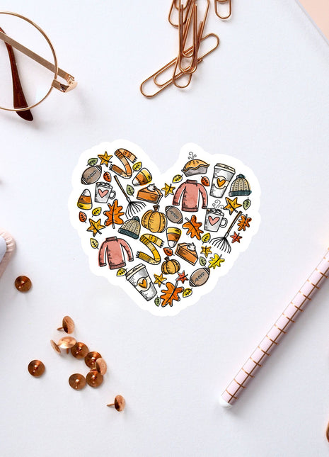 a heart-shaped sticker with various fall-themed items such as pumpkins, leaves, and a cup, surrounded by office supplies like pens, paper clips, and a notebook.