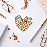 a heart-shaped sticker with various fall-themed items such as pumpkins, leaves, and a cup, surrounded by office supplies like pens, paper clips, and a notebook.