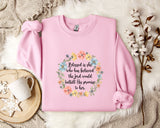 A pink sweatshirt with a floral wreath design and a quote printed on it, placed on a white fur rug.