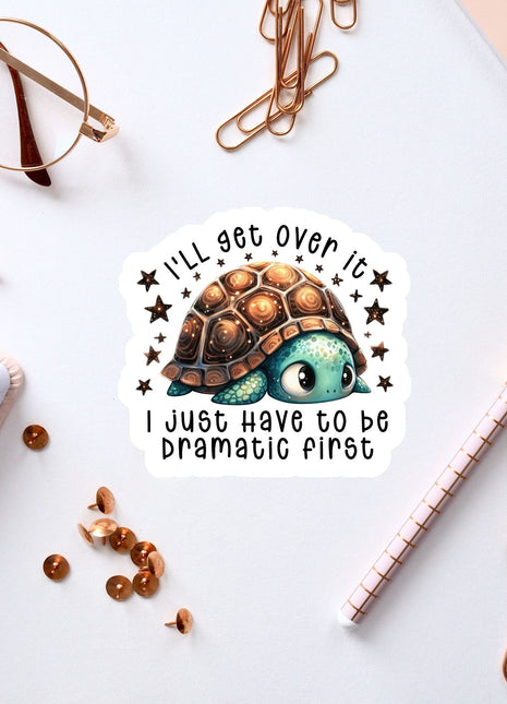 a sticker with a cartoon turtle and the text "I'll get over it, I just have to be dramatic first" surrounded by office supplies such as a pen, a pair of glasses, and a notebook.