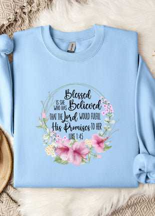 A light blue sweatshirt with a floral design and text printed on it, placed on a bed with a white blanket and a white mug.