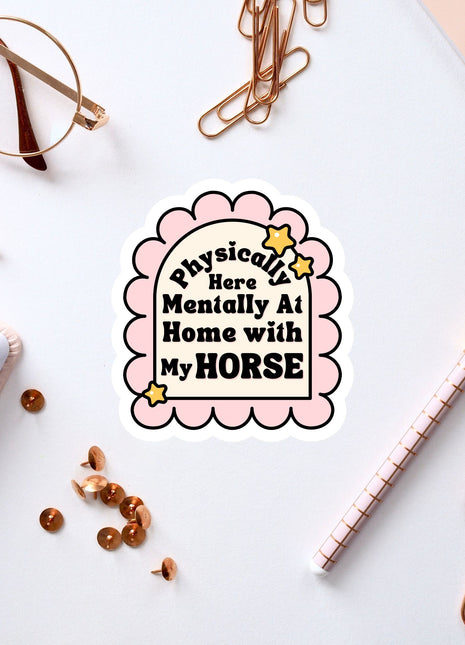 a sticker with a humorous text that reads "Physically Here Mentally At Home With My Horse" surrounded by various office supplies such as a pen, a notebook, and a pair of glasses.