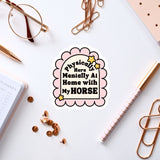 a sticker with a humorous text that reads "Physically Here Mentally At Home With My Horse" surrounded by various office supplies such as a pen, a notebook, and a pair of glasses.