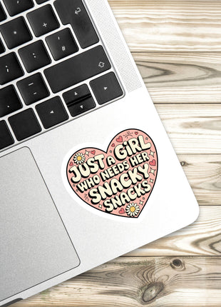 A laptop with a heart-shaped sticker on the palm rest, featuring a message about a girl who needs snacks.