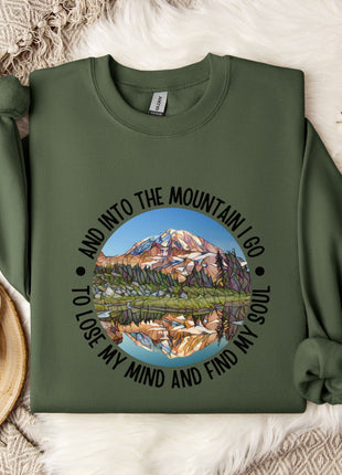 A green sweatshirt with a mountain landscape design and the text "And into the mountain I go, to lose my mind and find my soul" printed on it, along with a mug and some white flowers on a bed.