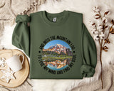 A green sweatshirt with a mountain landscape design and the text "And into the mountain I go, to lose my mind and find my soul" printed on it, along with a mug and some white flowers on a bed.
