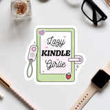 a sticker with the text "Lazy KINDLE Girlie" surrounded by various objects such as a pair of glasses, a watch, and a pen.