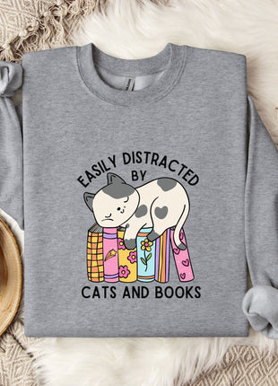 a gray sweatshirt with a graphic design of a cat sitting on a stack of books, accompanied by the text "EASILY DISTracted BY CATS AND BOOKS". The sweatshirt is laid out on a bed with a blanket and a mug, and there are some cotton balls and a branch of flowers nearby.
