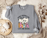 a gray sweatshirt with a graphic design of a cat sitting on a stack of books, accompanied by the text "EASILY DISTracted BY CATS AND BOOKS". The sweatshirt is laid out on a bed with a blanket and a mug, and there are some cotton balls and a branch of flowers nearby.