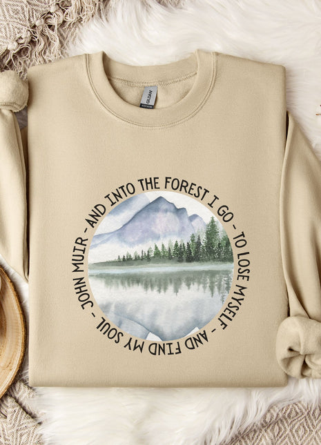 a beige sweatshirt with a circular design on it, depicting a mountain landscape with a lake and pine trees. The design also includes the text "John Muir - AND INTO THE FOREST I GO - TO LOSE MY SELF" around the edge of the circle. The sweatshirt is laid out on a bed with a white blanket, and there is a mug and some flowers nearby.
