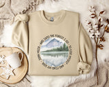 a beige sweatshirt with a circular design on it, depicting a mountain landscape with a lake and pine trees. The design also includes the text "John Muir - AND INTO THE FOREST I GO - TO LOSE MY SELF" around the edge of the circle. The sweatshirt is laid out on a bed with a white blanket, and there is a mug and some flowers nearby.