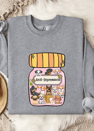 A gray sweatshirt with a graphic design of a jar filled with various cartoon animals, including dogs, cats, and birds, along with the text "Ani-Depressants" printed on it. The sweatshirt is laid out on a bed with a blanket and a mug, and there are some cotton balls and a branch of flowers nearby.