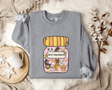 A gray sweatshirt with a graphic design of a jar filled with various cartoon animals, including dogs, cats, and birds, along with the text "Ani-Depressants" printed on it. The sweatshirt is laid out on a bed with a blanket and a mug, and there are some cotton balls and a branch of flowers nearby.