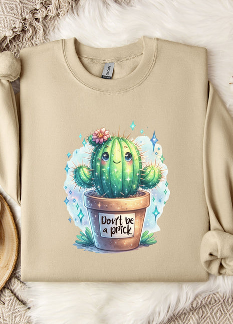 a beige sweater with a printed design of a cactus and the text "Don't be a prick" on it. The sweater is laid out on a bed with a white blanket and a white mug. There are also some white flowers and a white snowflake decoration in the scene.